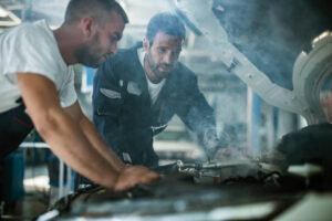 Auto mechanics cooperating while examining overheated car engine in auto repair shop. Focus is on man in blue uniform.